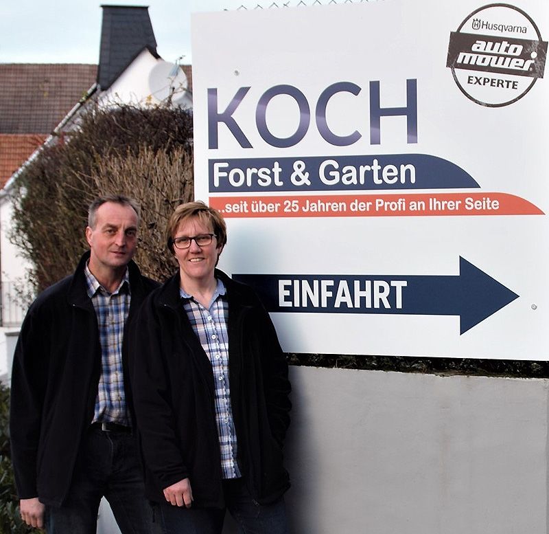 Eine Frau und ein Mann lächeln vor einem Werbeschild. Beide tragen karierte Hemden und dunkle Jacken.
