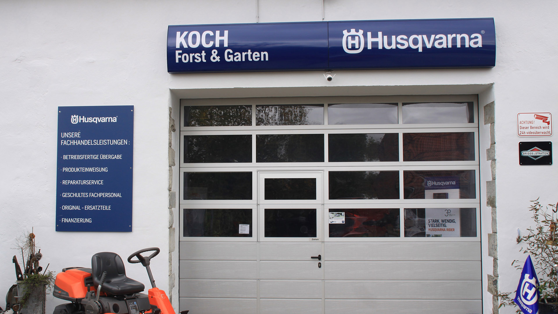 Geschäftseingang "Koch Forst & Garten", graues Rolltor, blaues Husqvarna-Logo, orangefarbener Rasenmäher.