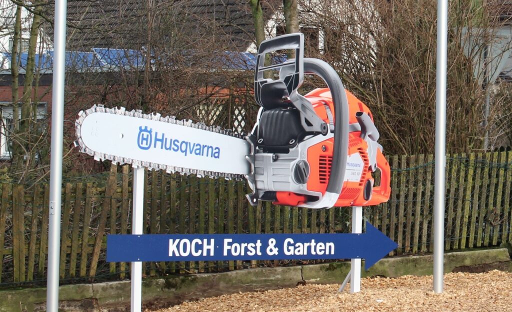 Großes, buntes Kettensägen-Modell mit "Husqvarna"-Logo auf einem Schild bei "KOCH Forst & Garten".