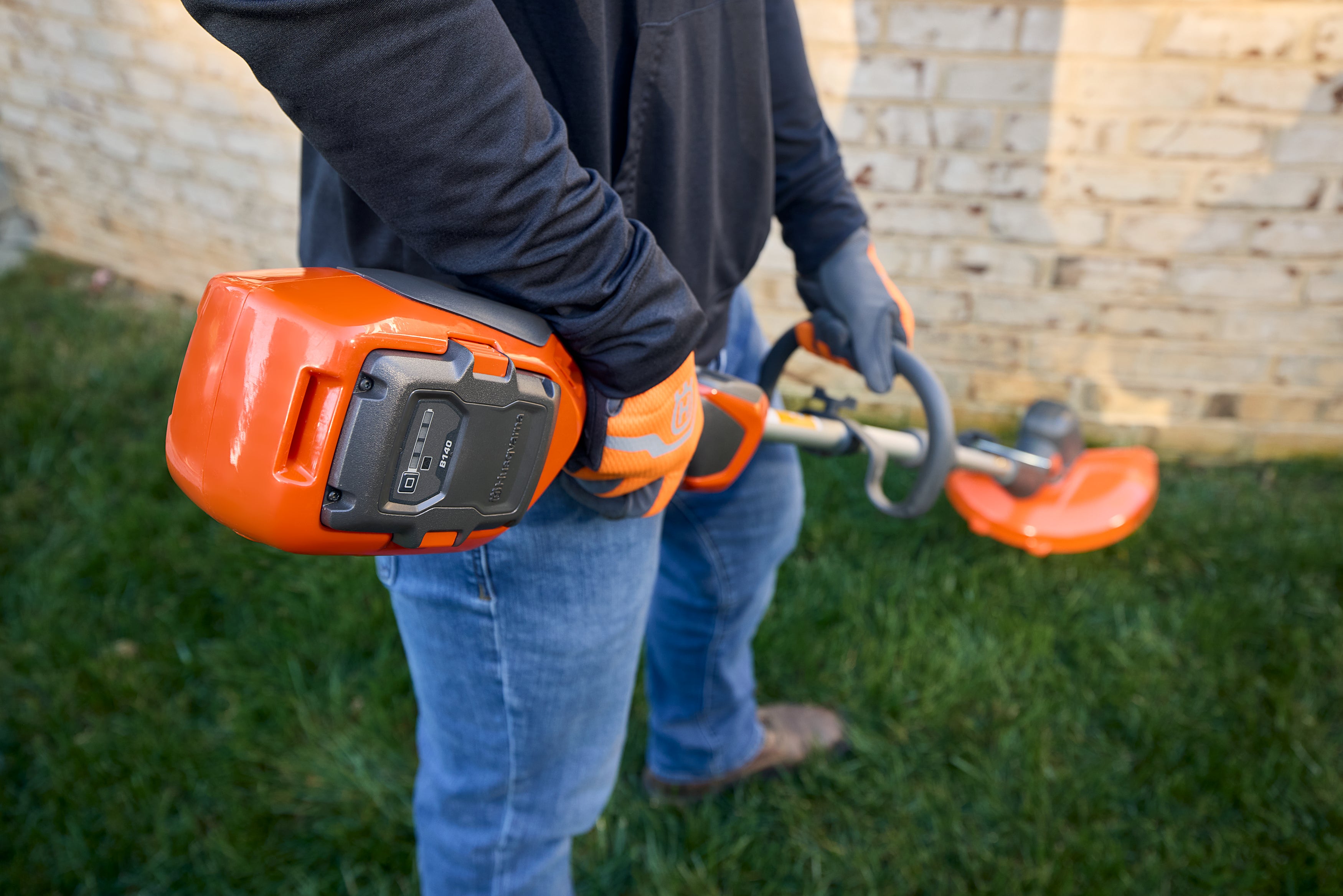 Person mit Trimmer, schwarze Kleidung, blaue Jeans, orangefarbene Akzente, steht auf Gras.