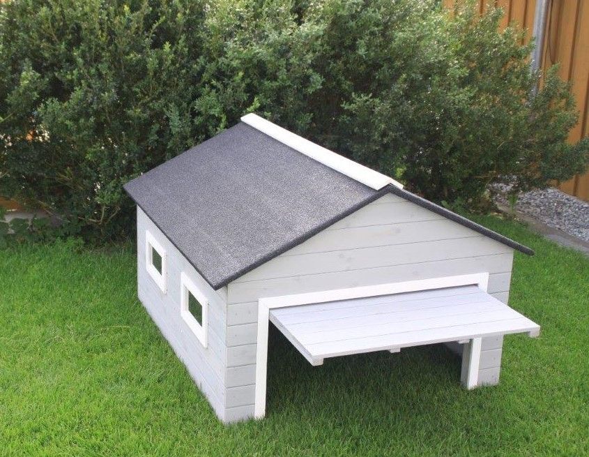 Ein graues Holz-Hundehaus mit schwarzem Dach auf grünem Rasen vor Büschen.
