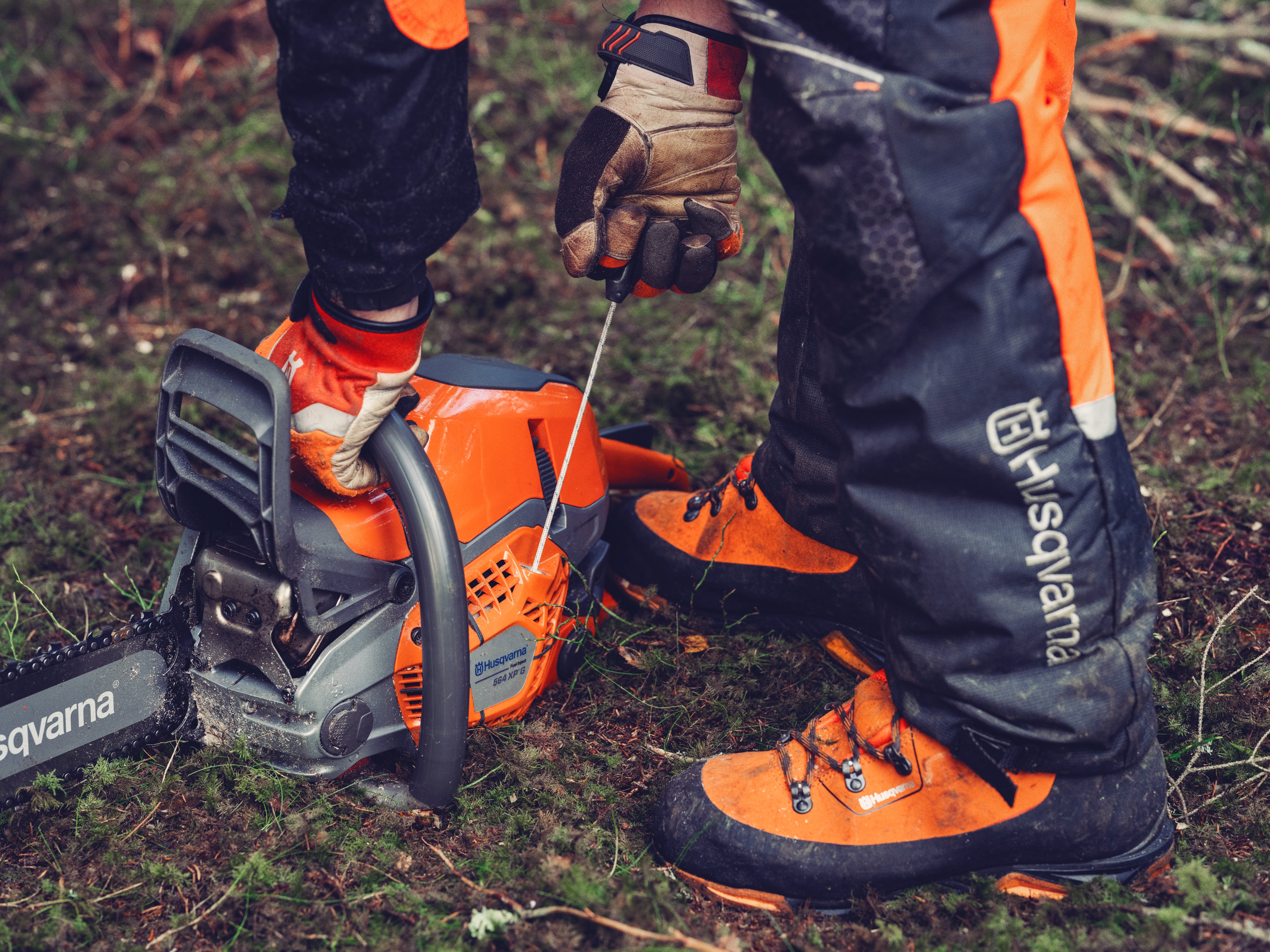 Person in Schutzkleidung zieht an einer orangefarbenen Motorsäge im Wald.